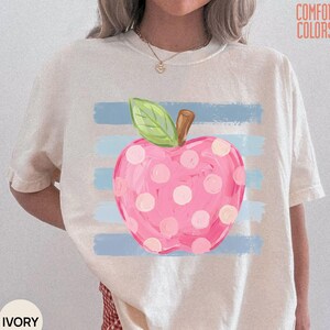 Pode incluir: Uma t-shirt de manga curta cor de marfim com um gráfico de uma maçã rosa com bolinhas brancas e uma folha verde. A maçã está centrada sobre riscas horizontais azul-claras. O texto COMFORT COLORS está impresso no canto superior direito.