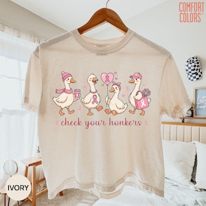 May include: An ivory t-shirt featuring a graphic of four white ducks wearing pink accessories and holding items like a coffee cup, a heart-shaped balloon, and a tote bag. The text "check your honkers" is printed in pink below the ducks.