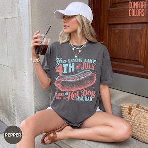 May include: A charcoal grey short-sleeve t-shirt featuring a graphic of a hot dog with the text "You look like the 4th of July makes me want a hot dog real bad" in red and light blue. The shirt is paired with a white baseball cap.