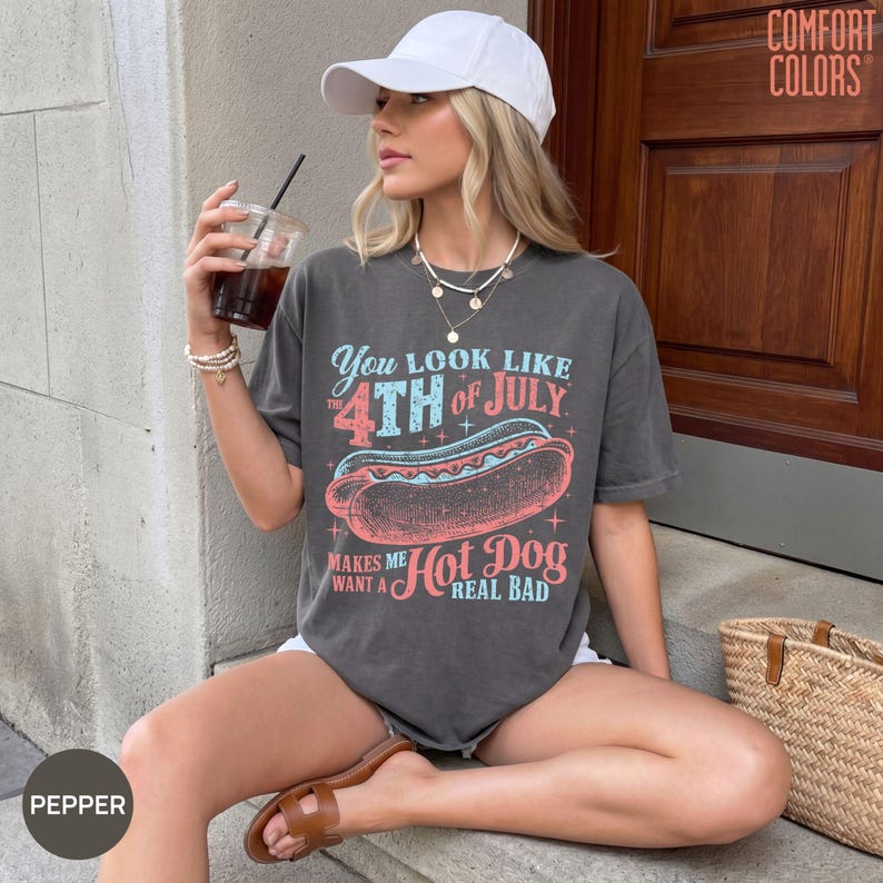 May include: A charcoal grey short-sleeve t-shirt featuring a graphic of a hot dog with the text "You look like the 4th of July makes me want a hot dog real bad" in red and light blue. The shirt is paired with a white baseball cap.