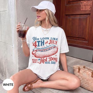 May include: A white short-sleeve t-shirt featuring a graphic of a hot dog and the text "You look like the 4th of July makes me want a hot dog real bad." The shirt has a crew neckline and a relaxed fit. A white baseball cap is also worn.