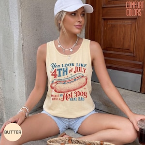 May include: A pale yellow sleeveless tank top featuring a graphic of a hot dog with red and blue text that reads: You look like 4th of July makes me want a hot dog real bad. The shirt has a crew neckline and a relaxed fit.