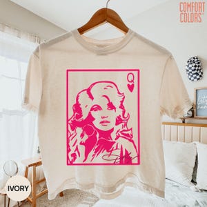May include: An ivory short-sleeve t-shirt featuring a hot pink graphic print of a woman with voluminous hair inside a rectangular frame. The design resembles a playing card with a heart symbol and the letter Q in the top right corner.