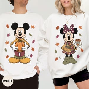May include: Two white crewneck sweatshirts featuring Disney characters. The left sweatshirt shows Mickey Mouse holding a pumpkin, while the right sweatshirt shows Minnie Mouse holding a coffee cup. Both designs include scattered autumn leaves.