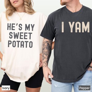 Könnte beinhalten: Ein elfenbeinfarbenes T-Shirt mit dem schwarzen Text HE'S MY SWEET POTATO und ein pfefferfarbenes T-Shirt mit dem beigen Text I YAM. Beide T-Shirts haben einen abgenutzten, strukturierten Schriftstil und kurze Ärmel als passendes Set.