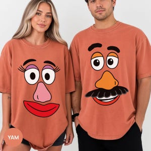 May include: Two terracotta-colored crew neck t-shirts featuring graphic prints of cartoon potato character faces. The left shirt displays a face with eyelashes, purple eyeshadow, a pink nose, and red lips. The right shirt shows a face with a mustache, orange nose, and a wide smile.