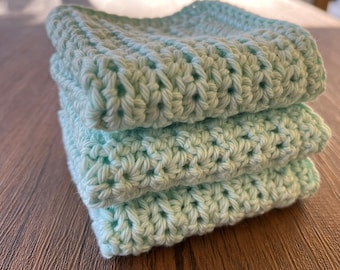 May include: A stack of four light green crocheted dishcloths. The dishcloths are made with a simple stitch pattern and have a soft, absorbent texture.