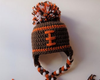May include: A brown and orange crocheted hat with a football design. The hat features a pom-pom on top with white, brown, and orange yarn. The ear flaps are braided with brown, white, and orange yarn, with tassels at the ends.