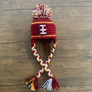 Peut inclure: Chapeau crocheté rouge et or avec un pompon blanc et des liens tressés blancs et or. Le chapeau a un motif de football blanc sur le devant.