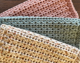 May include: Three crocheted dishcloths in different colours: beige with speckles of orange, green, and a light pink. The dishcloths are stacked on top of each other.