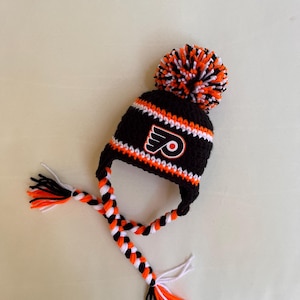 Könnte beinhalten: Eine gehäkelte Wintermütze in Schwarz, Orange und Weiß mit Ohrenklappen und einem Bommel. Die Mütze zeigt das Logo der Philadelphia Flyers auf der Vorderseite. Die Ohrenklappen sind mit orangefarbenem, schwarzem und weißem Garn geflochten.