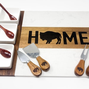 May include: A rectangular serving board with white marble and dark wood sections, featuring the text HOME with a buffalo silhouette. Includes three white square bowls with dark wooden spoons, and four cheese knives with wooden handles and buffalo icons.