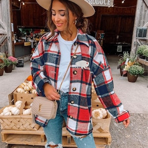 May include: A red, white, and navy blue plaid button-down shirt jacket featuring a small white buffalo patch on the chest, worn over a white t-shirt with blue distressed denim jeans, a beige fedora hat, and a beige quilted crossbody bag.