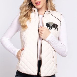 May include: A cream-colored quilted vest featuring a stand-up collar, a full-length front zipper, and dark brown trim. An embroidered patch of a black and grey buffalo is positioned on the upper left chest. The vest includes two side pockets.