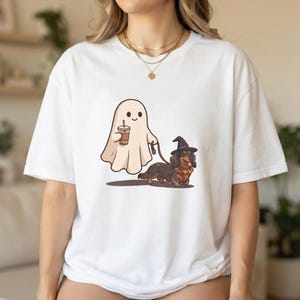 May include: A white short-sleeved t-shirt featuring a graphic print of a cartoon ghost holding a coffee cup and walking a long-haired dachshund wearing a black witch hat. The ghost is off-white and the dog is brown and black with a collar.