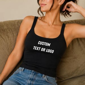May include: A black tank top featuring the white text CUSTOM TEXT OR LOGO printed on the chest. The garment has a scoop neckline and wide shoulder straps, paired with blue denim jeans. The design is simple, focusing on the customizable area.