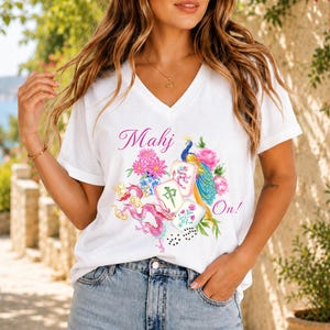 May include: A white V-neck t-shirt featuring a colorful graphic print with the text Mahj On! The design includes a peacock, a pink dragon, mahjong tiles, and floral elements in shades of pink, blue, green, and yellow on the front of the shirt.