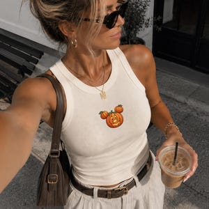 May include: A cream-colored ribbed tank top featuring a small embroidered graphic of three orange pumpkins arranged in a circular shape, paired with light-colored trousers, a dark brown leather belt, and a matching dark brown shoulder bag.