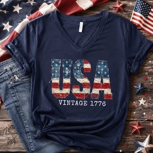 May include: Navy blue V-neck t-shirt with the word "USA" in a distressed American flag design. The text "VINTAGE 1776" is below the word "USA". The shirt is paired with blue jeans and patriotic decorations.