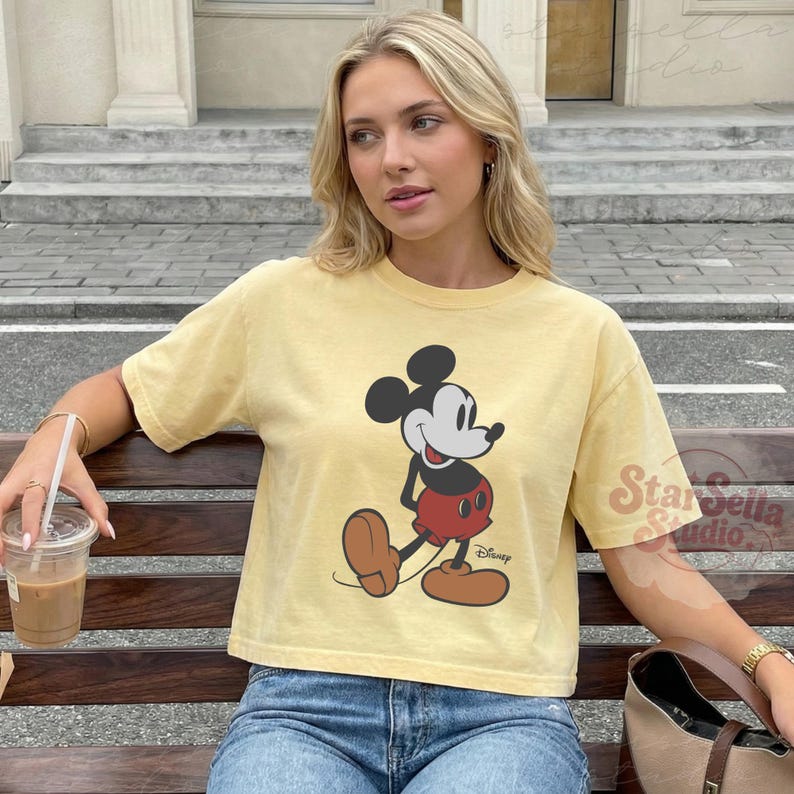 May include: A pale yellow t-shirt with a Mickey Mouse design. The cartoon character is in black, red, and brown. The shirt is worn with blue jeans. The Disney logo is printed on the shirt.