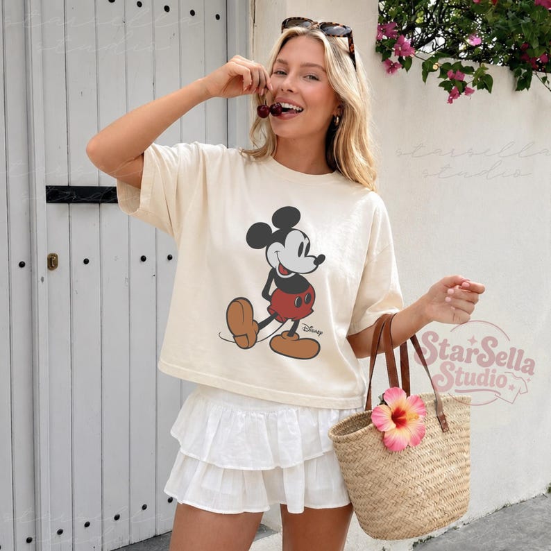 May include: Cream-coloured t-shirt with a Mickey Mouse graphic in black, red, and brown. The shirt is worn with a white ruffled skirt and a woven straw tote bag with a pink flower.