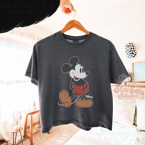 May include: A dark grey t-shirt with a Mickey Mouse design. The cartoon mouse is wearing red shorts and brown shoes. The shirt is displayed on a wooden hanger. The Disney logo is visible.