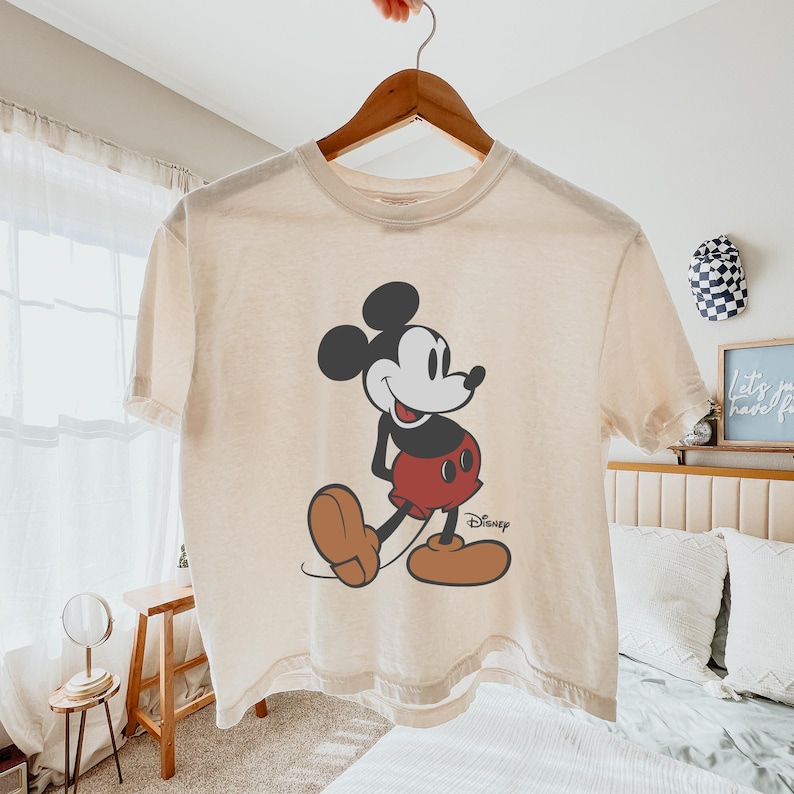 May include: Cream-coloured t-shirt featuring a classic Mickey Mouse graphic. The cartoon character is depicted in black, red, and brown, with the Disney logo printed below. The shirt is hanging on a wooden hanger.