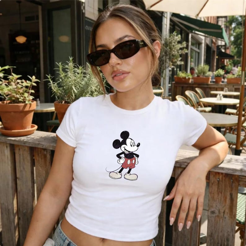 May include: A white short-sleeved t-shirt featuring a centered graphic of Mickey Mouse. The character is depicted in a classic style with black ears, a white face, and red shorts. The shirt has a crew neckline and a fitted silhouette, providing a clean look.