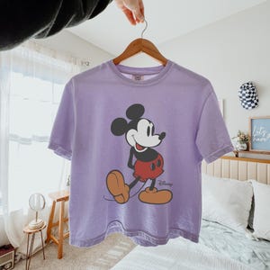 May include: A lilac t-shirt with a Mickey Mouse graphic. The cartoon character is in black, red, and tan, with the Disney logo. The shirt is displayed on a wooden hanger. The t-shirt is a casual wear item.