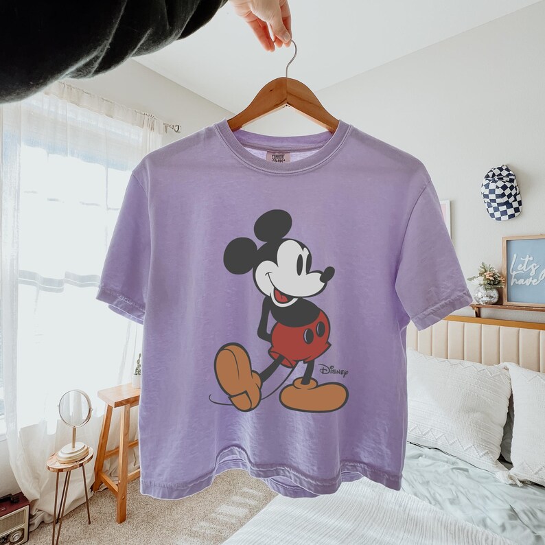 May include: A lilac t-shirt with a Mickey Mouse graphic. The cartoon character is in black, red, and tan, with the Disney logo. The shirt is displayed on a wooden hanger. The t-shirt is a casual wear item.