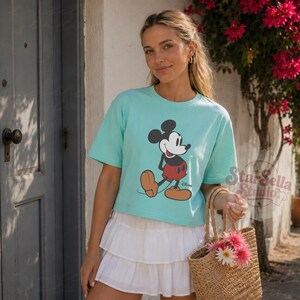 May include: A light blue t-shirt with a Mickey Mouse graphic. The shirt is worn with a white tiered skirt and a woven tote bag with pink flowers. The image is taken outside.
