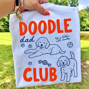 以下が含まれることがあります： オレンジ色の文字でDOODLE DAD CLUBとプリントされた白いTシャツ。デザインには、2匹のドゥードル犬、足跡、ボールの紺色の線画と、life is better with doodlesという文字が含まれています。Tシャツは緑の背景を背にして掲げられています。