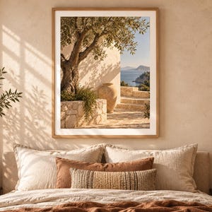 Op de afbeelding: Een ingelijste kunstdruk met een mediterrane scène met een olijfboom, een stenen plantenbak, een grote terracotta amfora en uitzicht op zee. Het kunstwerk hangt boven een bed met beige en bruine kussens en een textuurrijke deken.
