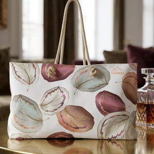 Puede incluir: Un bolso tote blanco con un patrón repetitivo de hojas estilizadas en tonos rosa empolvado, verde salvia y cobre. El bolso tiene dos asas gruesas de cuerda color crema anudadas a través de ojales metálicos y el texto SAKAKEKA impreso.
