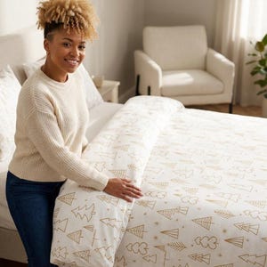 Puede incluir: Una funda nórdica blanca con funda de almohada a juego que presenta un patrón repetitivo de árboles de Navidad dibujados con líneas doradas y pequeños detalles de estrellas. La ropa de cama es de tela suave y tiene un diseño festivo.