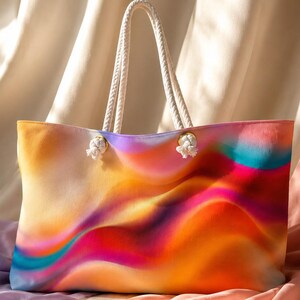 Puede incluir: Un bolso tote con un vibrante patrón de degradado multicolor con ondas fluidas en naranja, amarillo, rosa y turquesa. El bolso incluye dos asas gruesas de cuerda trenzada blanca que están aseguradas con ojales de metal en tono dorado.