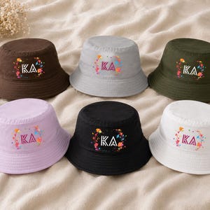 Puede incluir: Seis sombreros de pescador en varios colores: marrón, gris, verde, lavanda, negro y blanco. Cada sombrero presenta las letras "KA" en un diseño floral colorido. Los sombreros se muestran sobre una superficie de tono neutro.