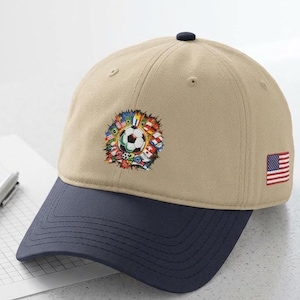Puede incluir: Una gorra de béisbol de color beige con visera azul oscuro y un botón en la parte superior. El frente presenta un balón de fútbol bordado rodeado de varias banderas internacionales y el texto 2020. Una pequeña bandera de EE. UU. está bordada a la derecha.