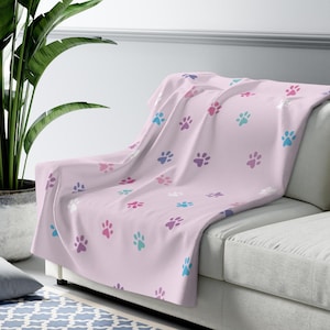 Puede incluir: Una manta de color rosa claro con un patrón repetitivo de pequeñas huellas de animales en varios colores, incluyendo azul, morado, blanco y rosa oscuro. La manta está colocada sobre un sofá, mostrando una textura suave y un diseño uniforme.