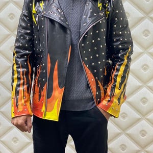 Peut inclure: Veste en cuir noire ornée de clous argentés, d'éclairs jaunes et de motifs de flammes jaunes, oranges et rouges sur les manches. La veste a un style motard classique avec un col cranté et une fermeture éclair.