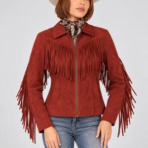 May include: A rust-colored suede jacket featuring long fringe detailing across the chest and down the length of both sleeves. The jacket includes a pointed collar, a central metal zipper closure, and is worn over blue denim jeans.