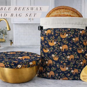 May include: Reusable beeswax bread bag set. The bag and bowl cover feature a navy blue fabric with a pattern of sloths and flowers. The bread bag holds a loaf of bread. The bowl cover sits on a gold bowl. The set is labeled "Beeswax Infused".
