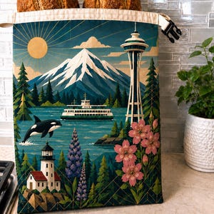 May include: A fabric bread bag with a roll-top closure and black buckle, featuring a printed illustration of a Pacific Northwest landscape with a mountain, the Space Needle, a ferry, an orca, a lighthouse, lupine flowers, and cherry blossoms.