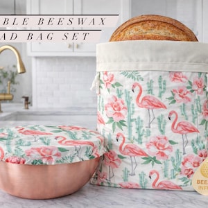 May include: A reusable beeswax bread bag set with a flamingo and floral print. The set includes a bread bag with a loaf of bread inside and a bowl cover. The bag and cover are white with pink flamingos, pink flowers, and green cacti.