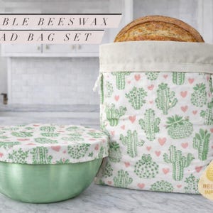 May include: A reusable beeswax bread bag set. The set includes a bread bag and a bowl cover, both with a white background and a pattern of green cacti and pink hearts. The bread bag is open, with a loaf of bread inside.