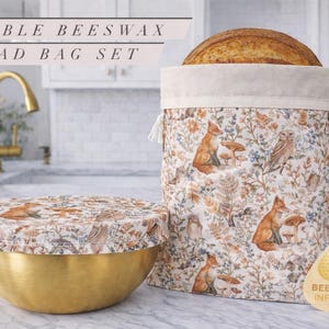 May include: A reusable beeswax bread bag set. The set includes a bread bag and a bowl cover, both featuring a woodland fox and mushroom pattern. The bag holds a loaf of bread. The text "REUSABLE BEESWAX BREAD BAG SET" is at the top of the image.