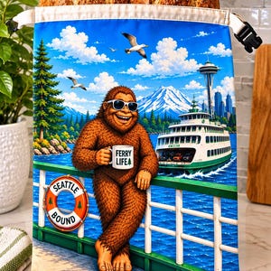May include: A fabric bread bag featuring a colorful illustration of a brown Bigfoot wearing sunglasses and holding a mug that says FERRY LIFE. The scene includes a ferry boat, the Space Needle, a mountain, and a life ring labeled SEATTLE BOUND.
