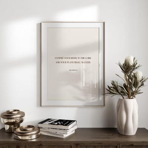 Puede incluir: Impresión enmarcada con el texto "COMMIT YOUR DEEDS TO THE LORD, AND YOUR PLANS SHALL SUCCEED". La impresión es de color beige claro, enmarcada en un marco plateado. Un jarrón blanco con flores y objetos decorativos de metal están sobre una superficie de madera.