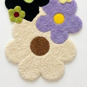 May include: A tufted rug shaped like a cluster of flowers with a large cream-colored bloom featuring a brown center, a purple bloom with a yellow center, a black bloom with a light green center, and a small green flower with a dark red center.