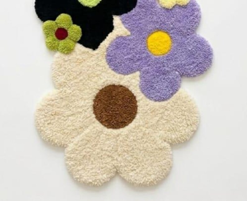 May include: A tufted rug shaped like a cluster of flowers with a large cream-colored bloom featuring a brown center, a purple bloom with a yellow center, a black bloom with a light green center, and a small green flower with a dark red center.